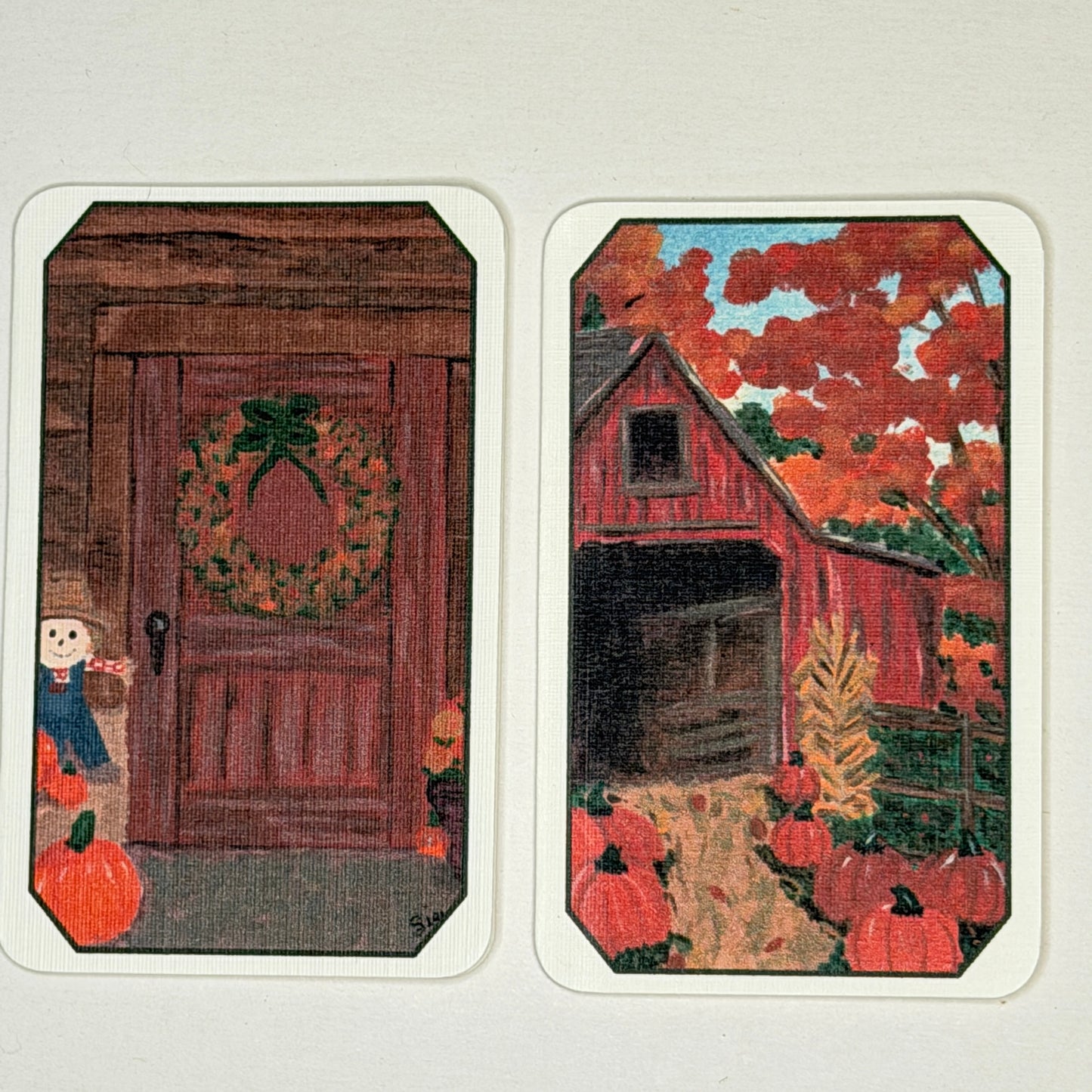 Two vintage autumn-themed cards or prints with rounded corners, displayed side-by-side on a light beige background. The left card shows a rustic brown door with a green and orange wreath, a small scarecrow, and pumpkins.