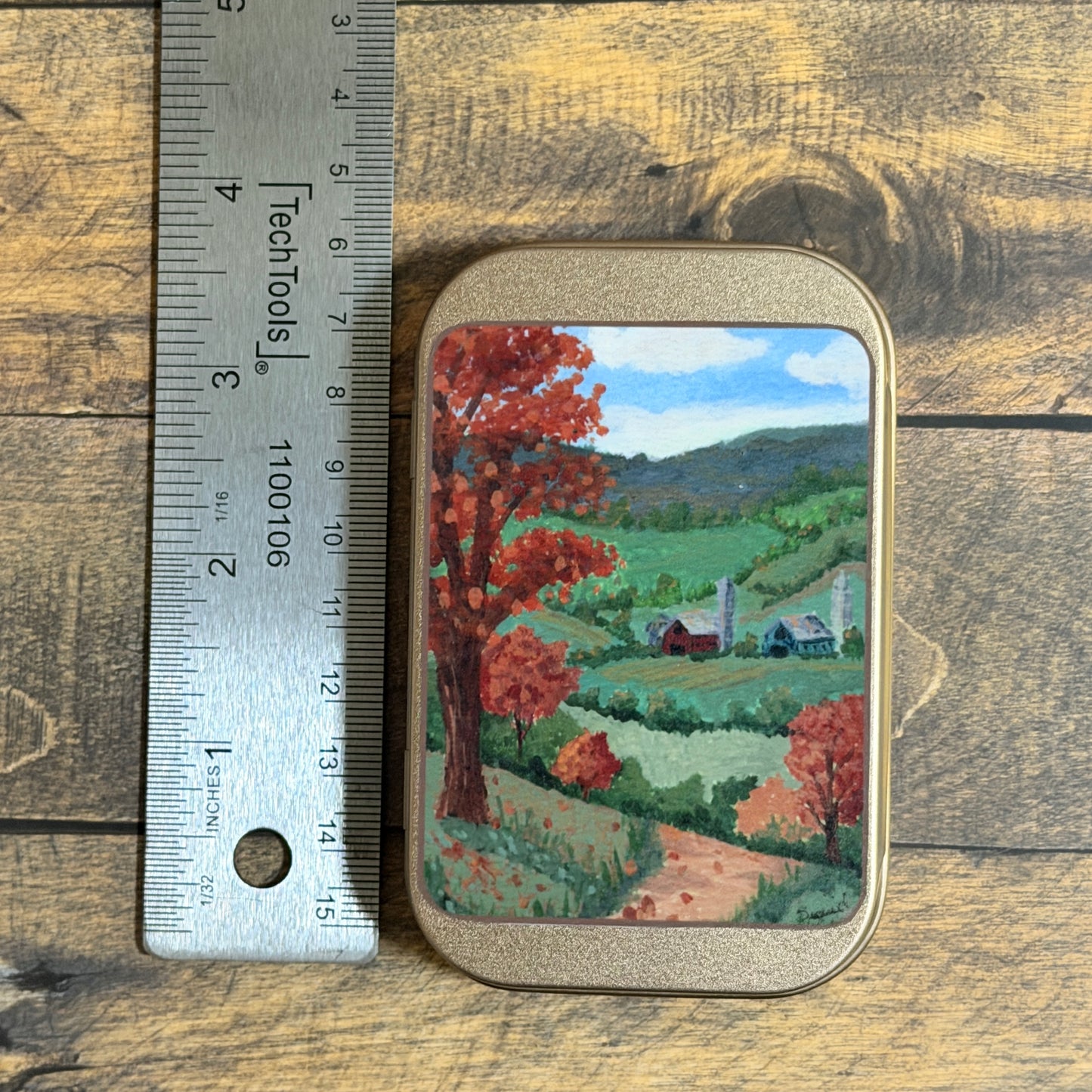 A small rectangular tin with rounded corners sits on a rustic wood surface next to a metal ruler for scale. The tin features a painted landscape scene with red autumn trees, green hills, blue sky, and small farm buildings.
