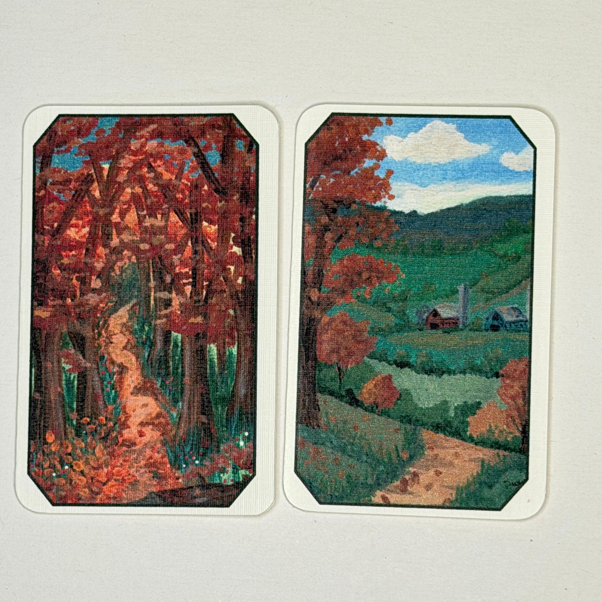 Two rectangular art cards with rounded corners displayed side by side on a light background. Each card shows a painted landscape scene: the left features a forest path with red autumn foliage, and the right shows a green valley with small buildings and a winding path under a blue sky.