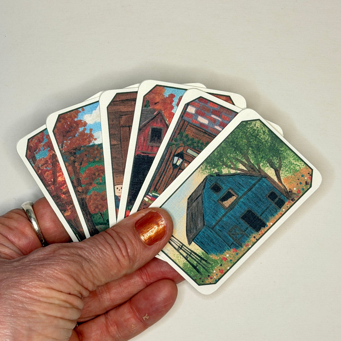 A hand holds a fan of illustrated cards showing rustic farmhouse scenes with autumn trees and a blue cabin, against a plain light background.