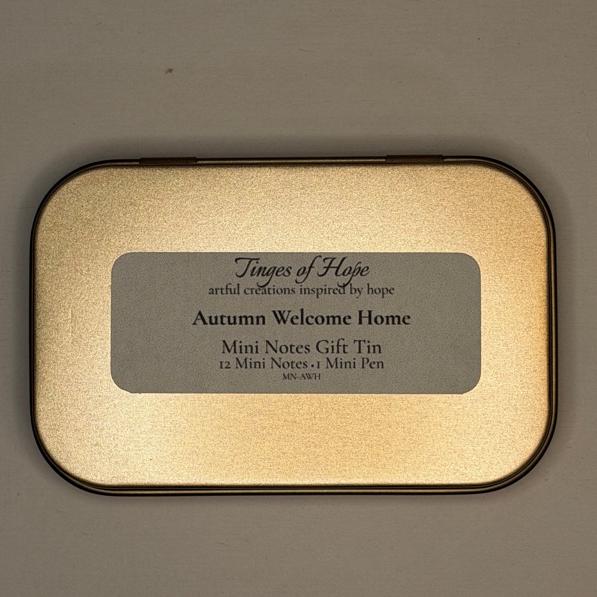 A rectangular gold-toned metal tin with rounded corners and a black border, featuring a gray label that reads 'Tinges of Hope' and 'Autumn Welcome Home Mini Notes Gift Tin'.