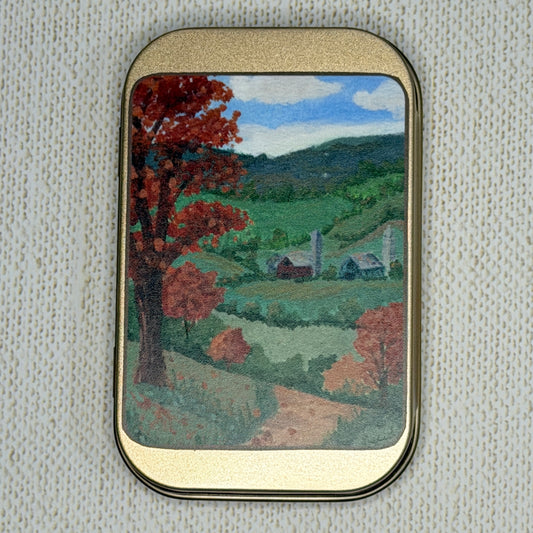 A rectangular rose gold metal tin with rounded corners, featuring a painted landscape scene of a rural valley with red trees, green hills, and a blue sky. The tin is set against a textured off-white fabric background.