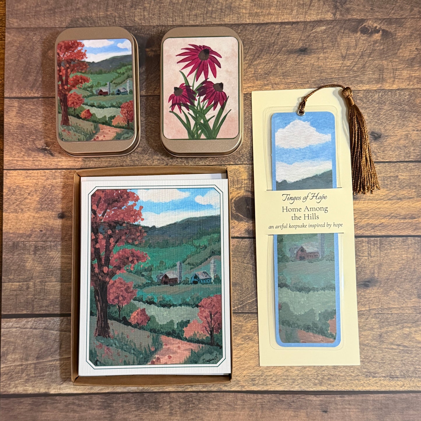 A coordinated set of home decor items featuring a landscape painting of 'Home Among the Hills.' The set includes two small metal tins, a boxed card or print, and a bookmark with a tassel, all arranged on a rustic wooden surface.