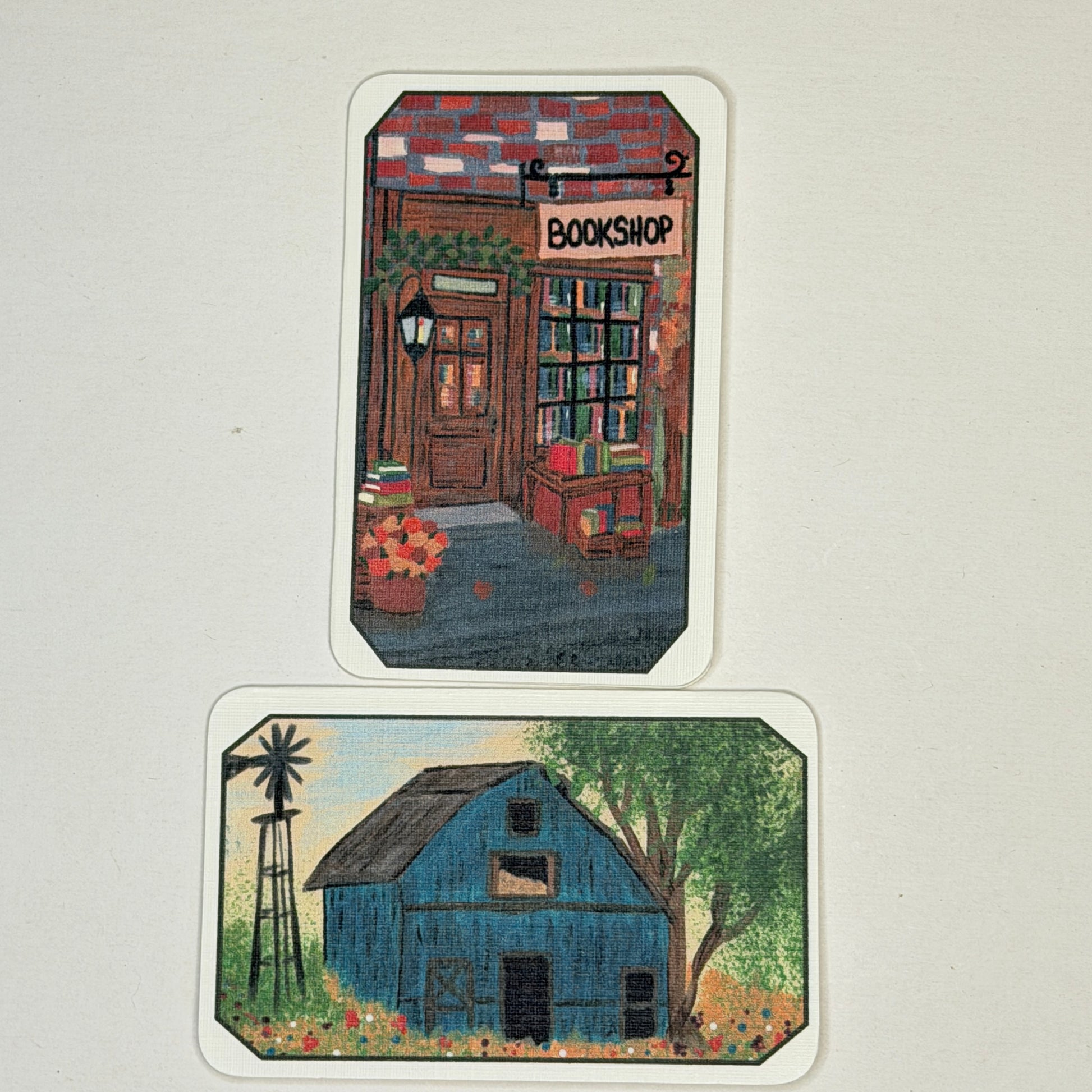 Two illustrated art cards with rounded corners displayed on a light beige background. The top card shows a vintage-style bookshop storefront with flowers and a sign reading BOOKSHOP.