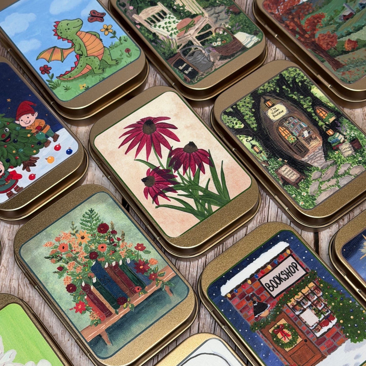 A collection of small rectangular metal tins with rounded corners, each decorated with a different whimsical illustrated scene. The designs include a green dragon, a cozy cabin, flowers, a treehouse, a bookshelf with books, a snowy bookshop, and other holiday and nature motifs.