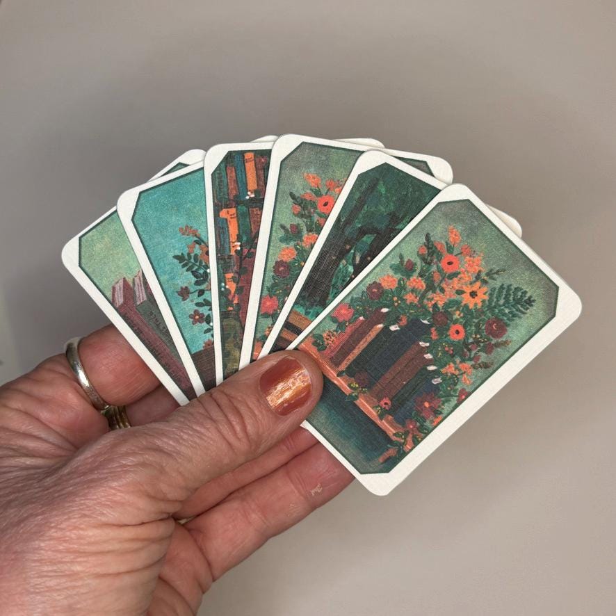 A hand holds a fan of illustrated oracle or tarot-style cards against a plain light gray background. The cards show floral and architectural scenes with teal, green, orange, and brown tones, and the hand has a gold ring and metallic nail polish.