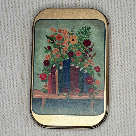 A rectangular rose gold tin with rounded corners, featuring a floral bouquet in red, orange, and green flowers set against a teal background, framed by a gold-toned metal border.
