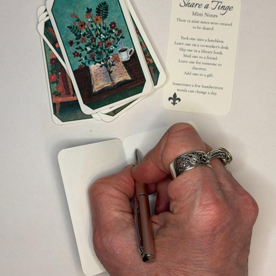 A hand holding a metallic pen over an open notebook, with a fanned deck of illustrated mini cards and a printed card titled 'Share a Tinge Mini Notes' on a white surface.