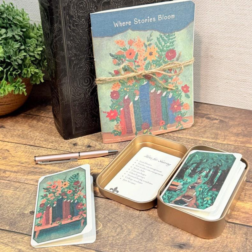 A boxed card or deck set titled "Where Stories Bloom" sits on a wooden table with its metal tin open, showing illustrated cards inside. A pen lies nearby, with a potted plant and a dark embossed book in the background.