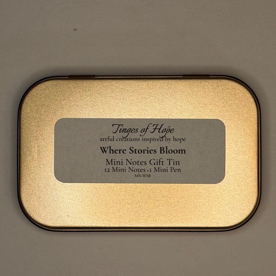 A square metallic gold tin with rounded corners and a black border, featuring a gray label that reads 'Tinges of Hoje' and 'Where Stories Bloom'.