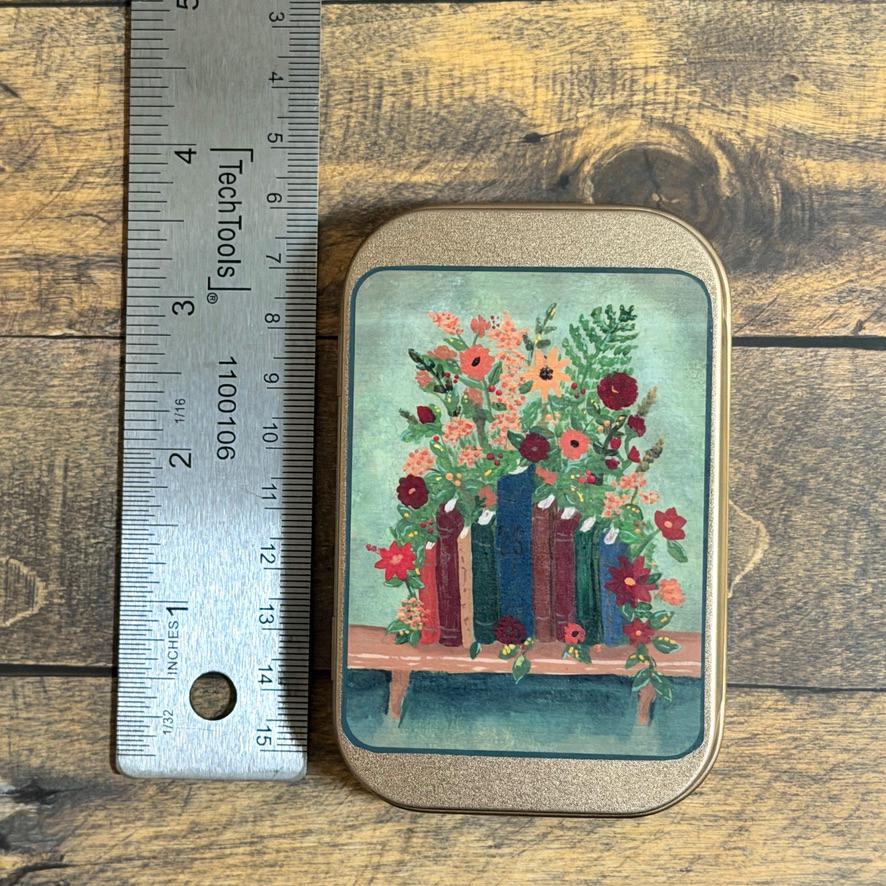 A small rectangular tin with rounded corners sits on a rustic wood surface next to a metal ruler for scale. The tin features an illustration of colorful flowers blooming from books against a light teal background.
